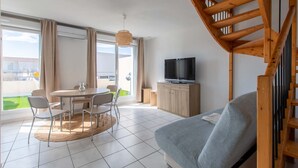 Un salon moderne comprenant un canapé, une table à manger avec des chaises, un téléviseur posé sur un meuble en bois et un escalier.