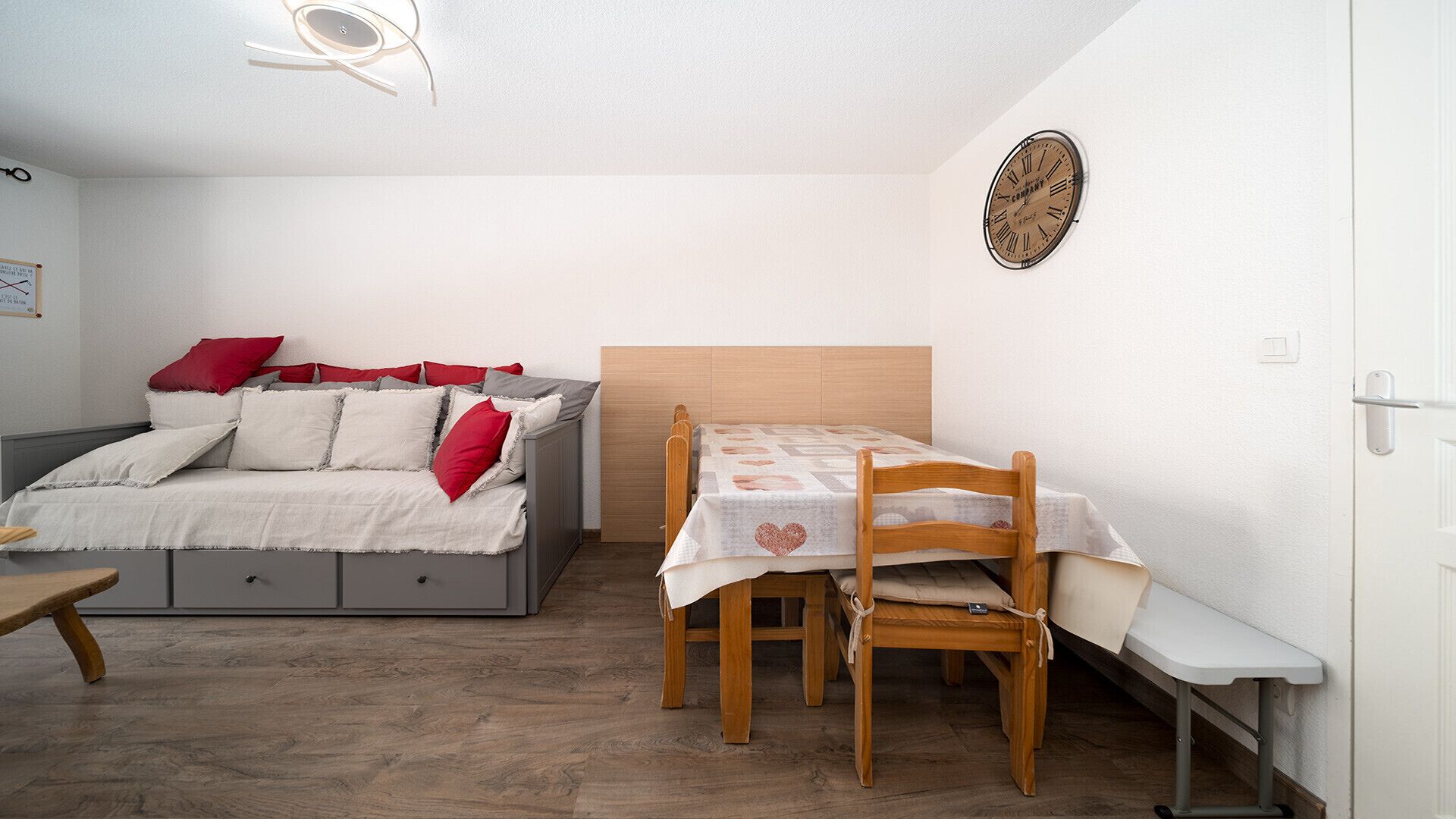 Ein kleiner, gut beleuchteter Raum mit einem Bett, einem Holztisch mit Stühlen und einer Wanduhr.