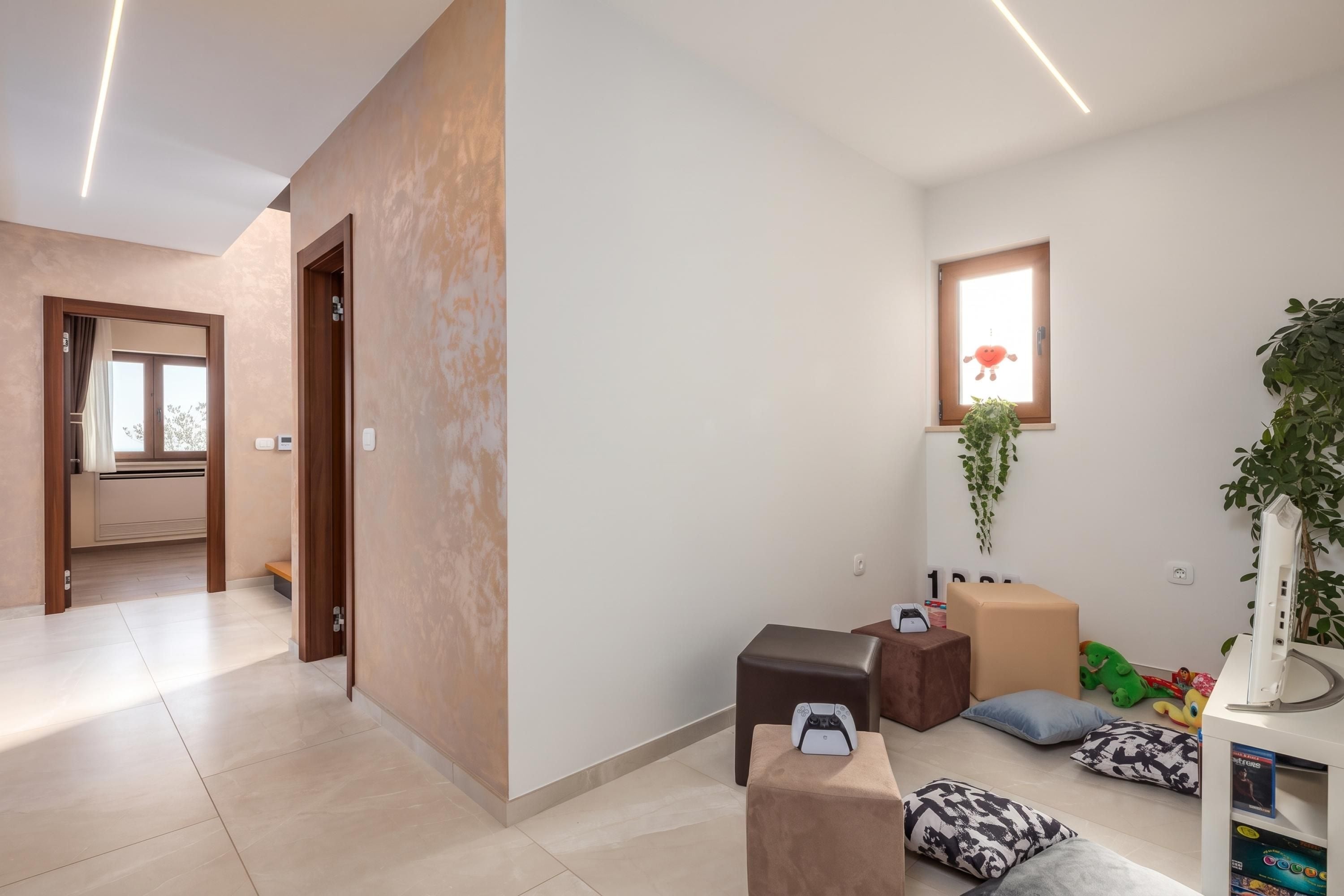 A modern hotel room with a hallway leading to a door, a small window with a plant, and a gaming console on a ottoman.