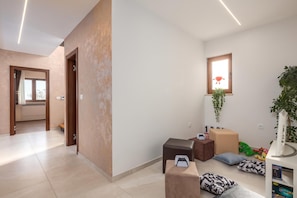 A modern hotel room with a hallway leading to a door, a small window with a plant, and a gaming console on a ottoman.