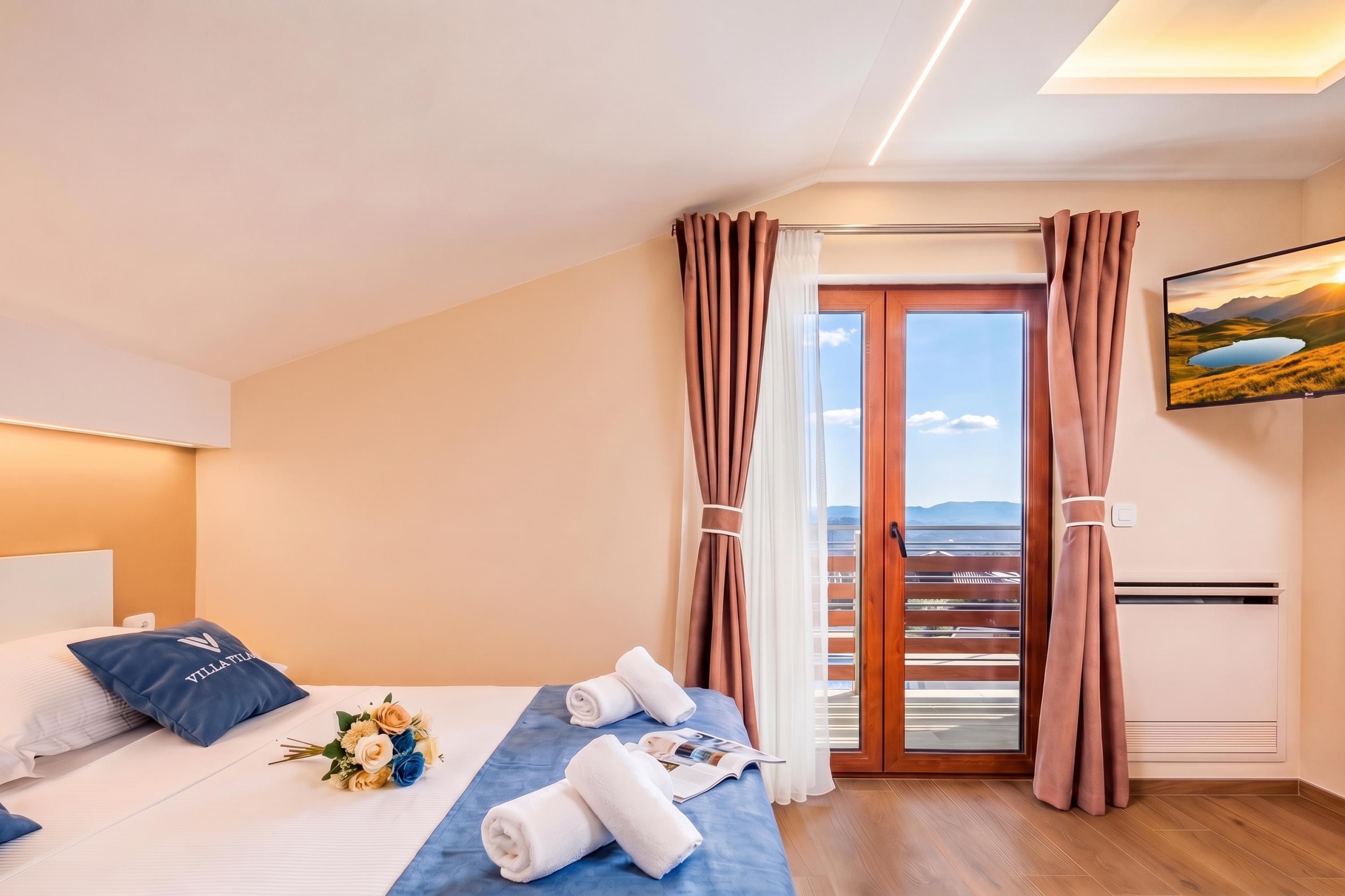 A hotel room with a bed, a blue bedspread, a bouquet of flowers, rolled towels, and a painting of a landscape.