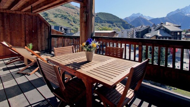 A wooden deck with a table and chairs, a potted plant, and a view of mountains and buildings. - Apt 4/6 ppl, terrace with a very nice view (Les Deux Alpes)