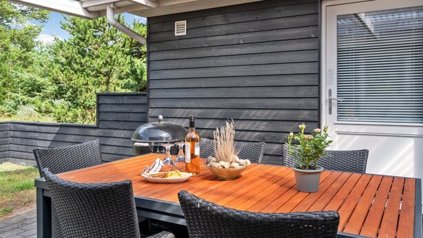 Een terras buiten met een houten tafel, rieten stoelen, een barbecue en een bloempot.