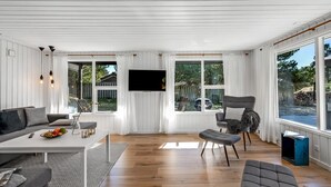 A modern living room with a sofa, armchair, and ottoman, a flat-screen TV mounted on the wall, and large windows offering a view of the outdoors. - "Allen" - 1km from the sea by Interhome (Blåvand)
