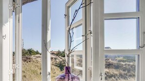 Ein Fenster mit Blick auf eine natürliche Landschaft, eine violette Vase mit Zweigen und ein klarer Himmel.