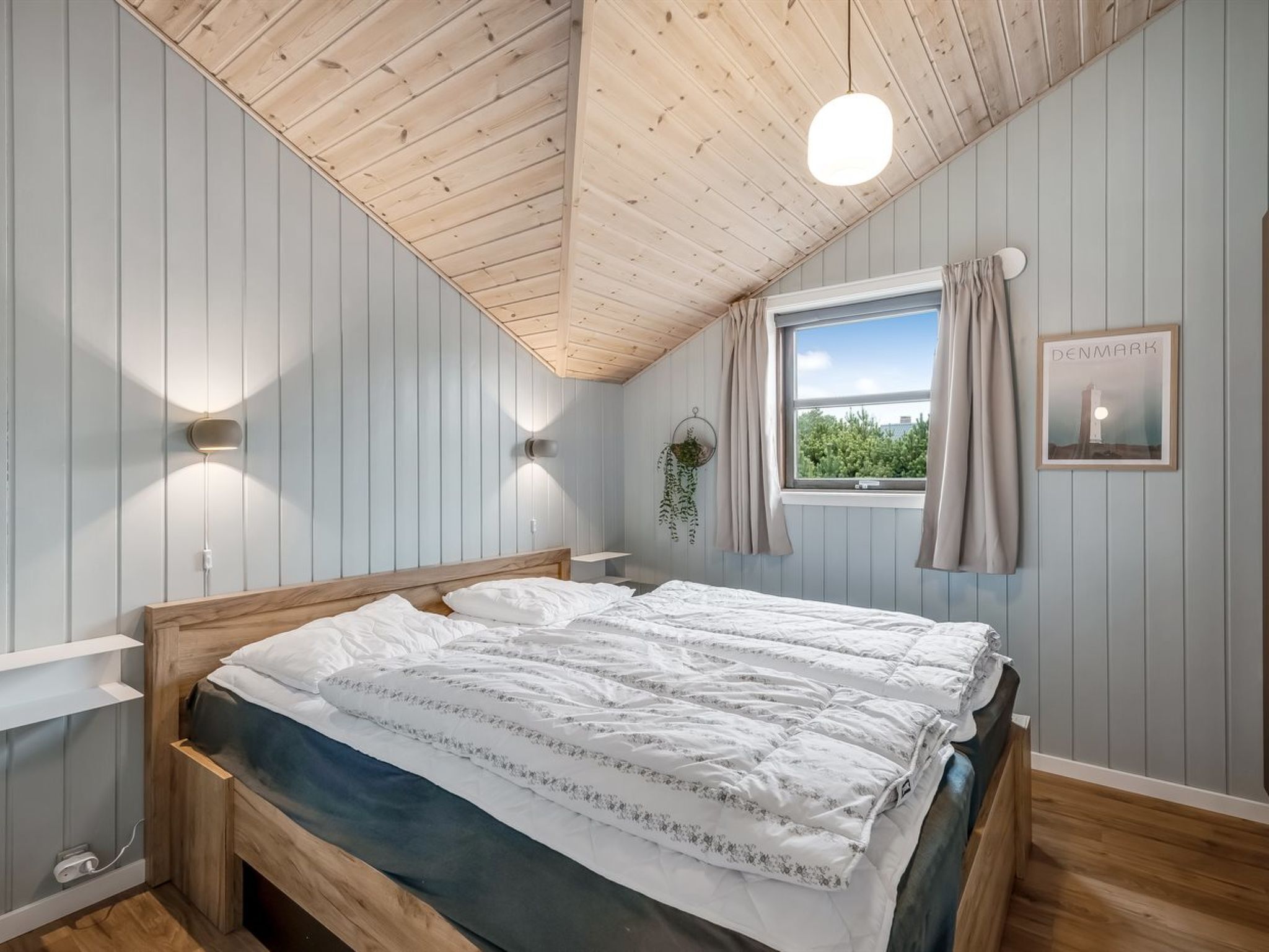 Une chambre avec un plafond en bois, un lit avec une literie blanche, une fenêtre avec des rideaux, un luminaire fixé au mur et un tableau représentant un phare.