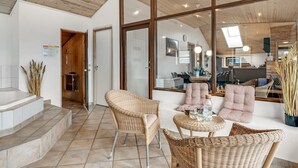 Ein geräumiger Wohnbereich mit Rattanmöbeln, eine durch große Fenster sichtbare Küche und ein modernes Design mit Holzbalkendecken.