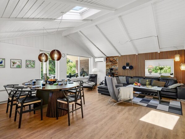 A spacious loft with a high ceiling, a dining area with a round table, a living area with a sofa and a fireplace, and a view of greenery outside.