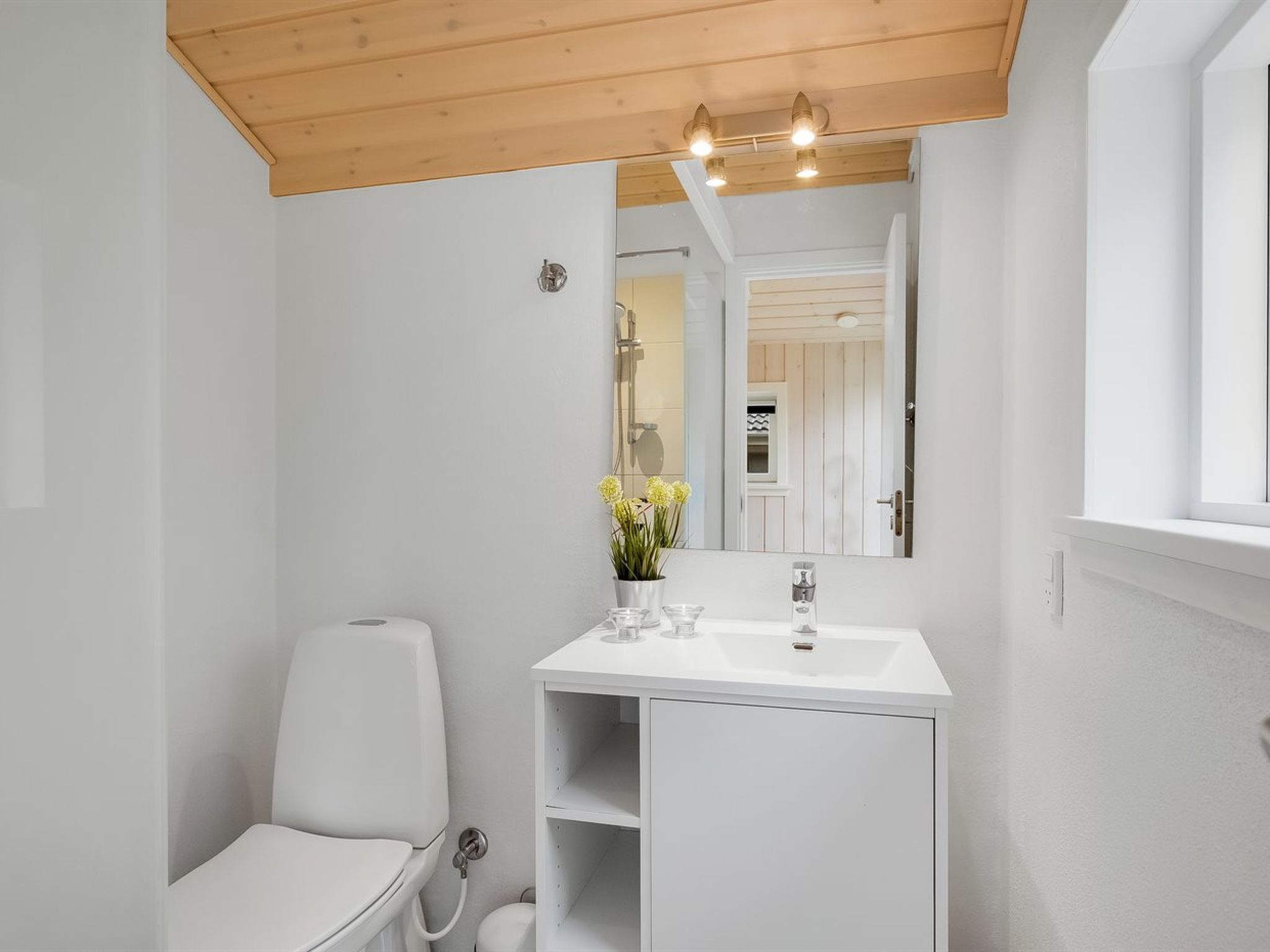 A modern bathroom with a white sink, mirror, and toilet. There is a wooden ceiling and a window on the right.