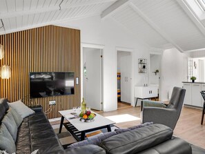 A modern living room with a large flat-screen TV, a wooden accent wall, a sofa, a coffee table with fruit, and a dining area with a white cabinet.
