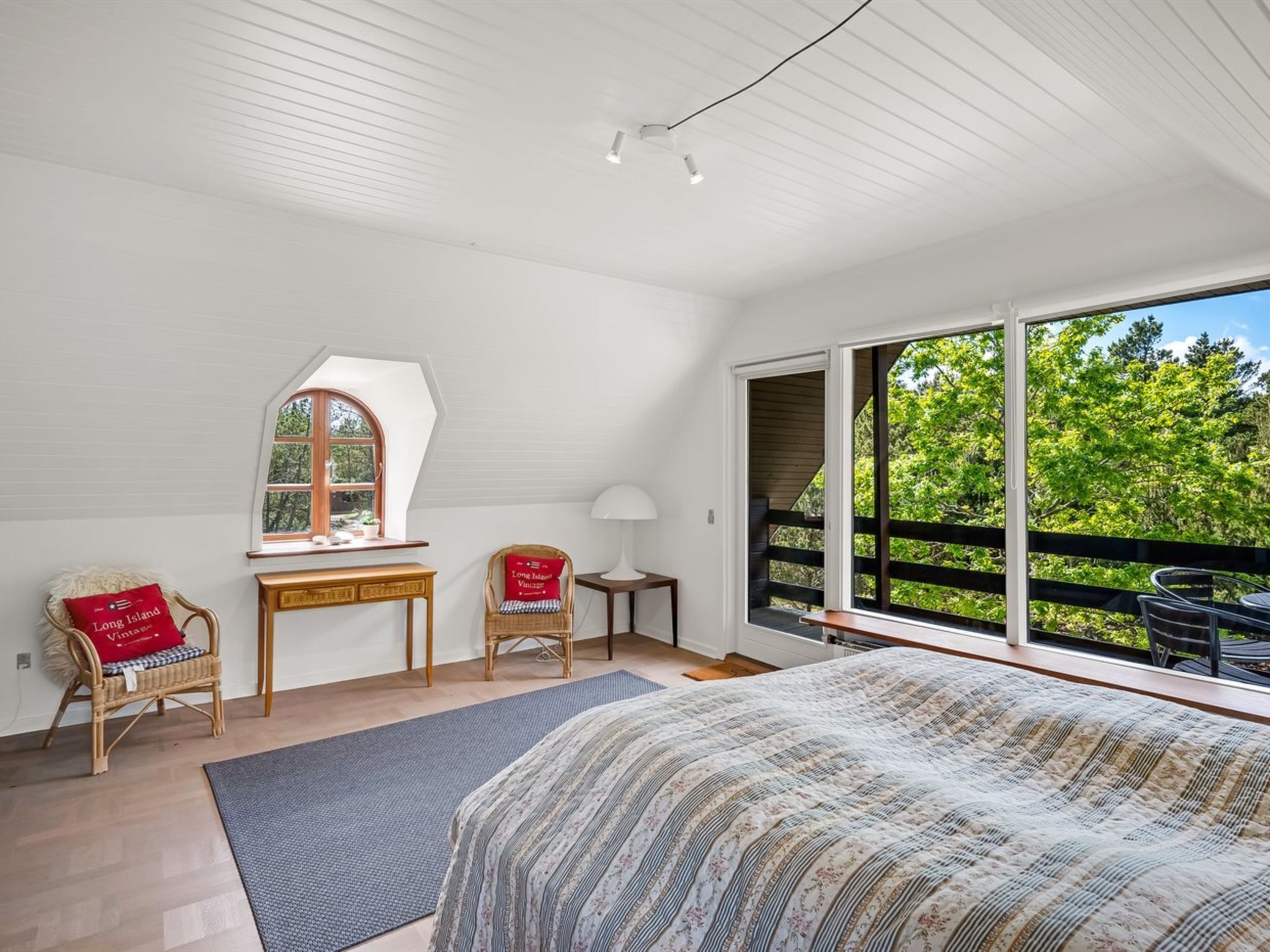 A bedroom with a large bed, a desk, two chairs, a window, and a view of greenery.