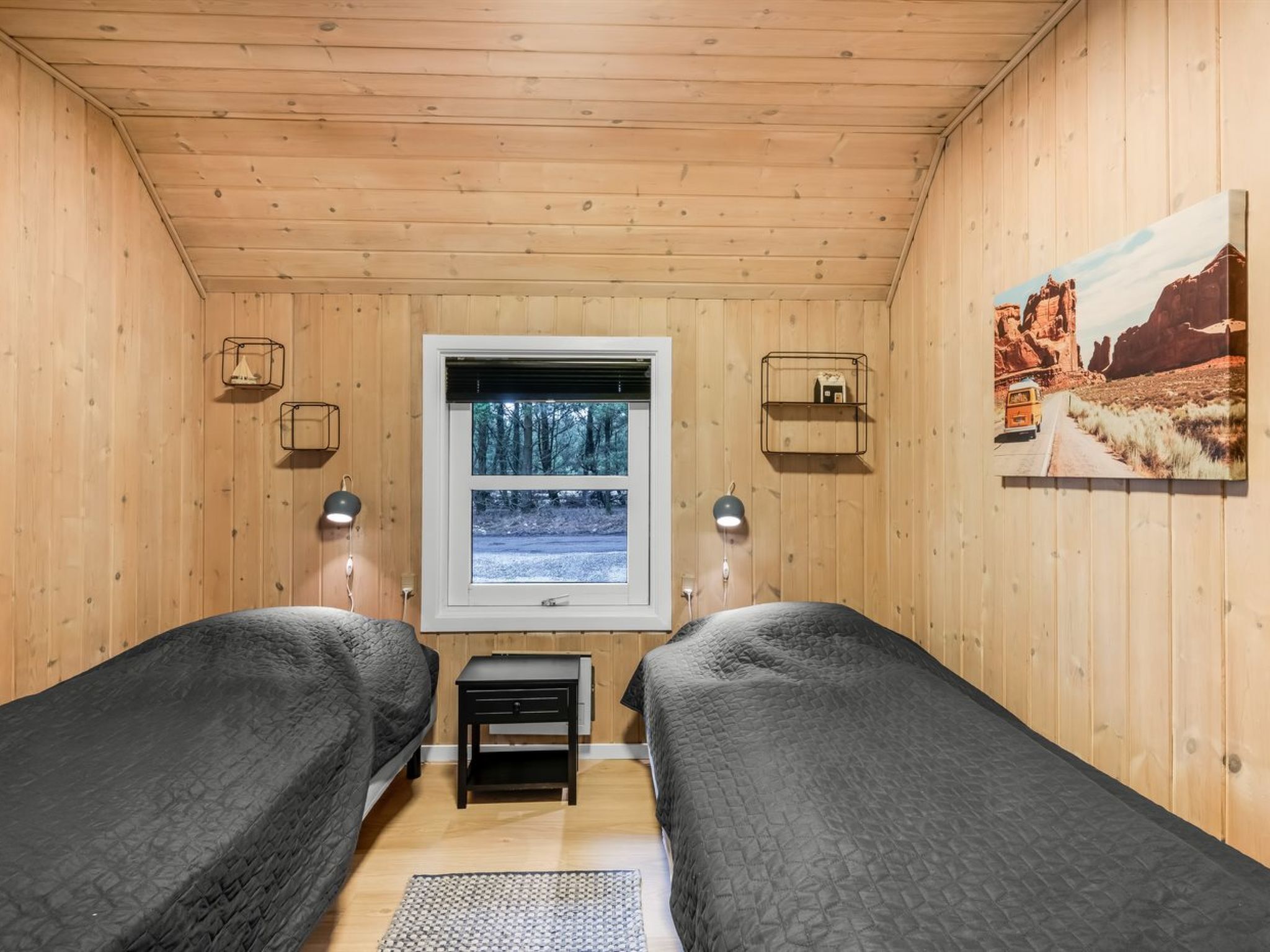 Una habitación tipo cabaña con dos camas, una ventana que da a árboles y un cuadro de un paisaje desértico.