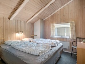 Ein Schlafzimmer mit Holzbalkendecke, einem Bett mit Duvet, einem Nachttisch, einem Sessel und einem Fenster mit Vorhängen.