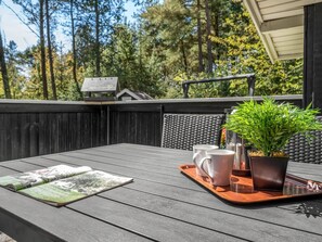 Eine Terrasse im Freien mit einem Holztisch, einer Ablage mit Tassen und einem Blumentopf, mit Bäumen im Hintergrund.