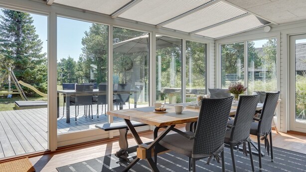 A sunroom with a wooden deck, large glass windows, and a dining area with chairs and a table.
