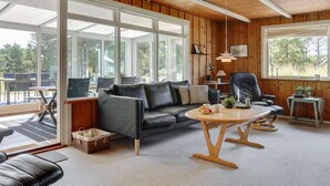 A cozy living room with a sofa, a round coffee table, and a leather chair. Large windows allow natural light and views of the outdoors.