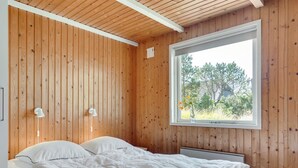 A bedroom with wooden panel walls, a large window with a view of trees, a bed with white bedding, and two bedside lamps.