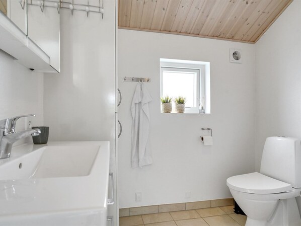 Un baño moderno con lavamanos, espejo y retrete blancos. Hay una ventana pequeña con plantas y una toalla colgada en la pared.