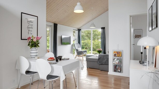 A modern living room with a dining area, featuring a white table, chairs, and a sofa. There is a large window providing natural light and a view of greenery outside. - "Jörgen" - 100m from the sea by Interhome (Esbjerg V)