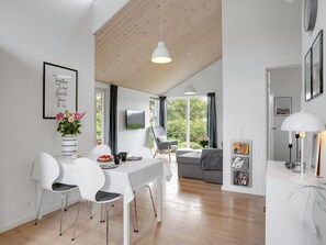 Un salon moderne avec un coin repas, comprenant une table blanche, des chaises et un canapé. Une grande fenêtre laisse entrer la lumière naturelle et offre une vue sur la verdure à l’extérieur.