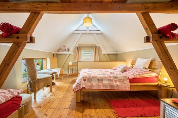 A cozy attic bedroom with a sloped ceiling, a bed with a quilt, a wicker chair, a small table, and a window with curtains.