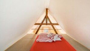 Un pequeño dormitorio abuhardillado con forma de triángulo, techo inclinado, una cama individual con colchón rojo y estructura de vigas de madera.