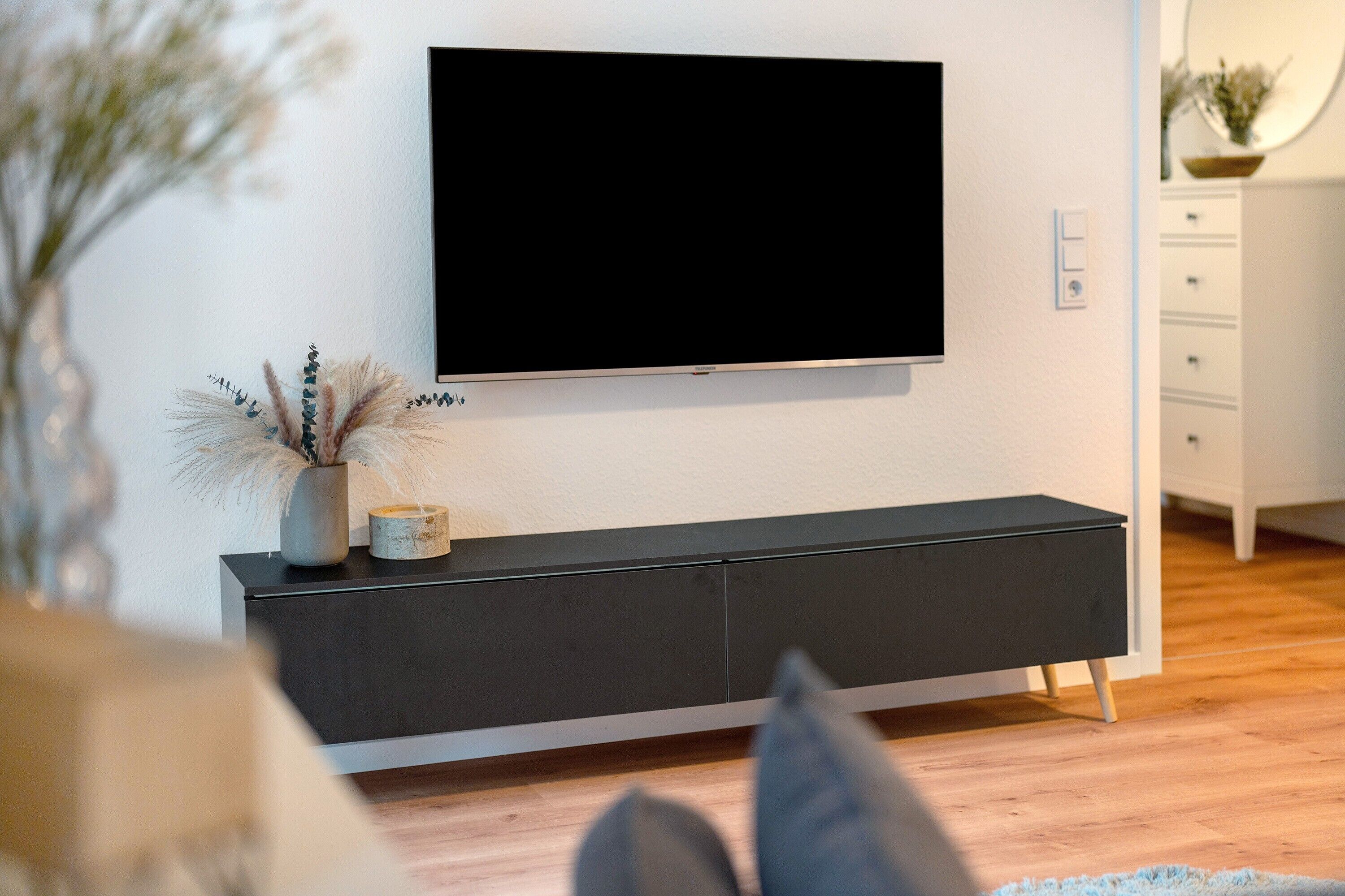 Una sala de estar moderna con un televisor de pantalla plana montado en una pared blanca, una consola oscura y suelo de madera.