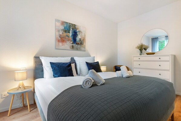 A neatly made bed with a quilted cover, a bedside lamp, a small wooden side table, a white dresser with a mirror, and a painting on the wall.