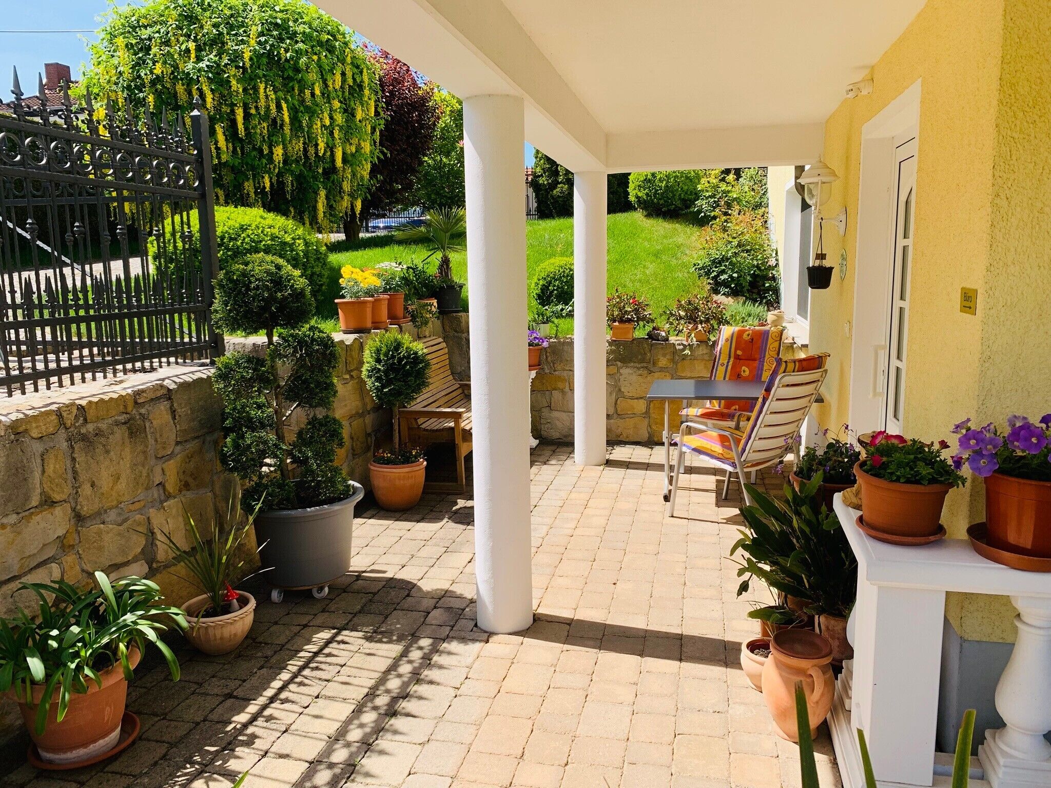 Ein überdachter Patio-Bereich mit Steinmauer, Kübelpflanzen, einer Holzbank und einem Tisch mit Stühlen.