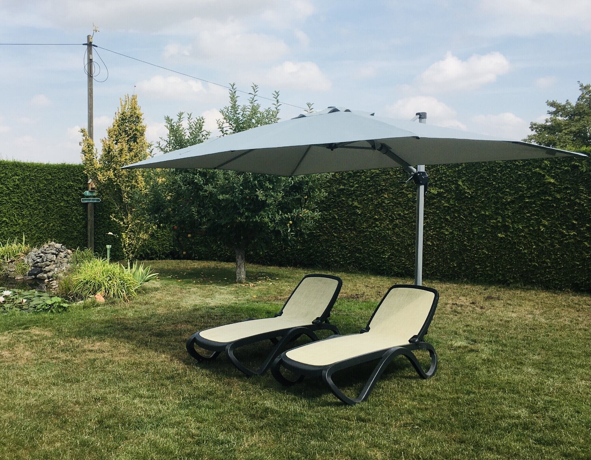 Zwei Liegestühle unter einem großen Sonnenschirm in einem grünen Garten mit einer Hecke und einem Vogelhäuschen.