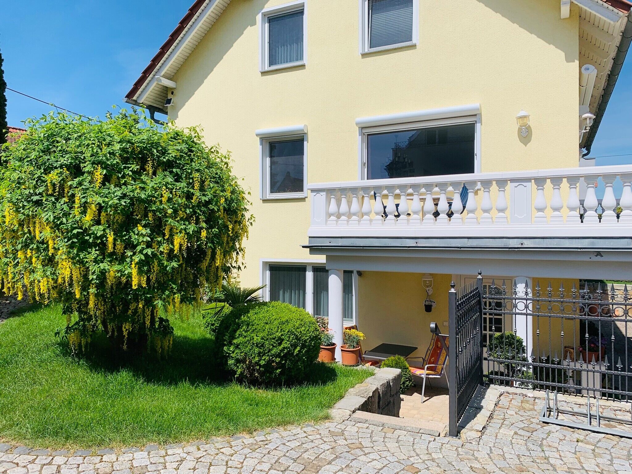 Ein zweistöckiges Gebäude mit einem Balkon, einem kleinen Garten mit gelben Blumen und einer gepflasterten Auffahrt.