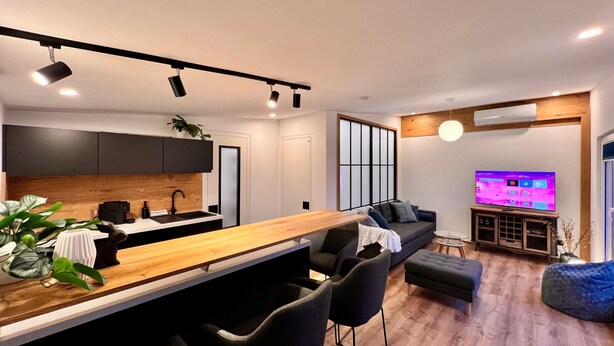 A modern kitchen with a wooden countertop and black chairs, a living area with a sofa and ottoman, and a TV displaying a streaming service.