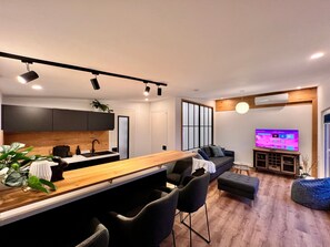 Una cocina moderna con encimera de madera y sillas negras, una sala de estar con sofá y reposapiés, y un televisor mostrando un servicio de streaming.