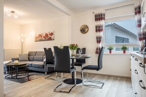 A modern living room with a sofa, dining table, chairs, and a large window with curtains. - Apartment 'Ferienhaus An Der Göhrde' with Private Terrace and Wi-Fi (Zernien)