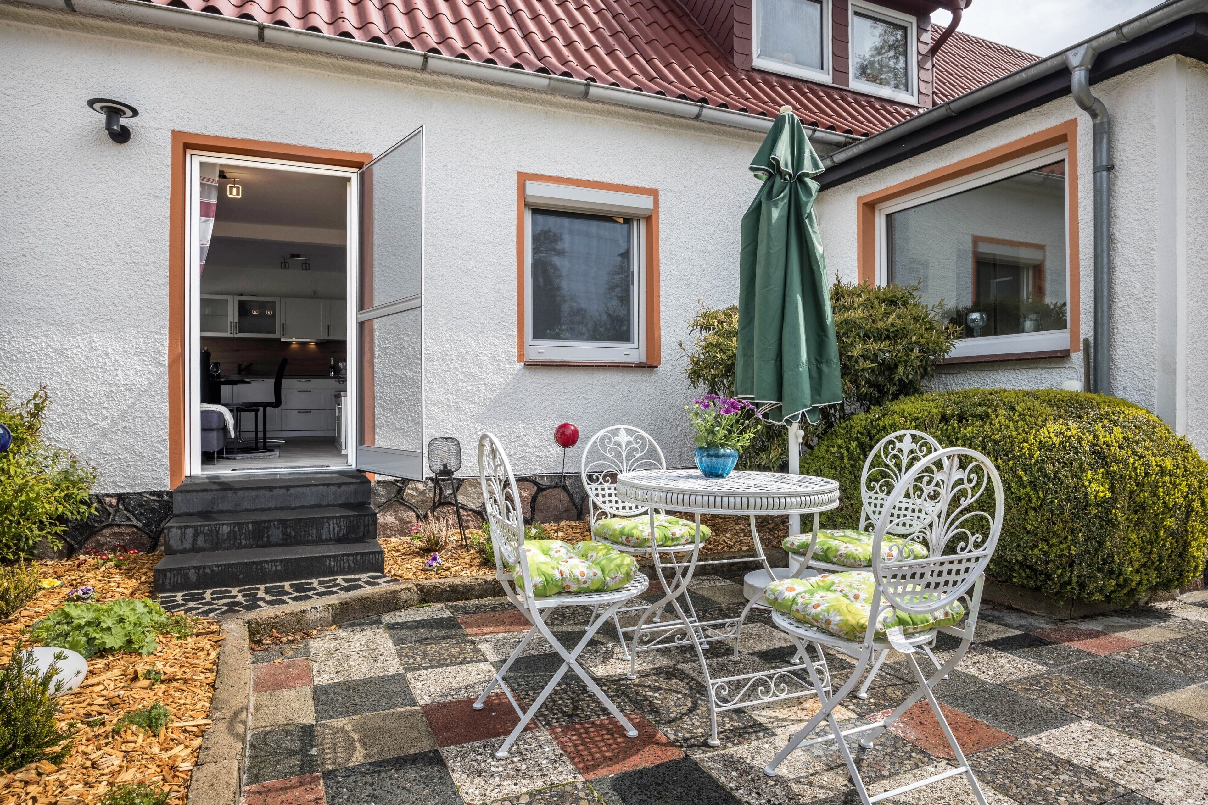 Una casa con un patio, una mesa de comedor con sillas, un paraguas verde y una ventana con marco blanco.