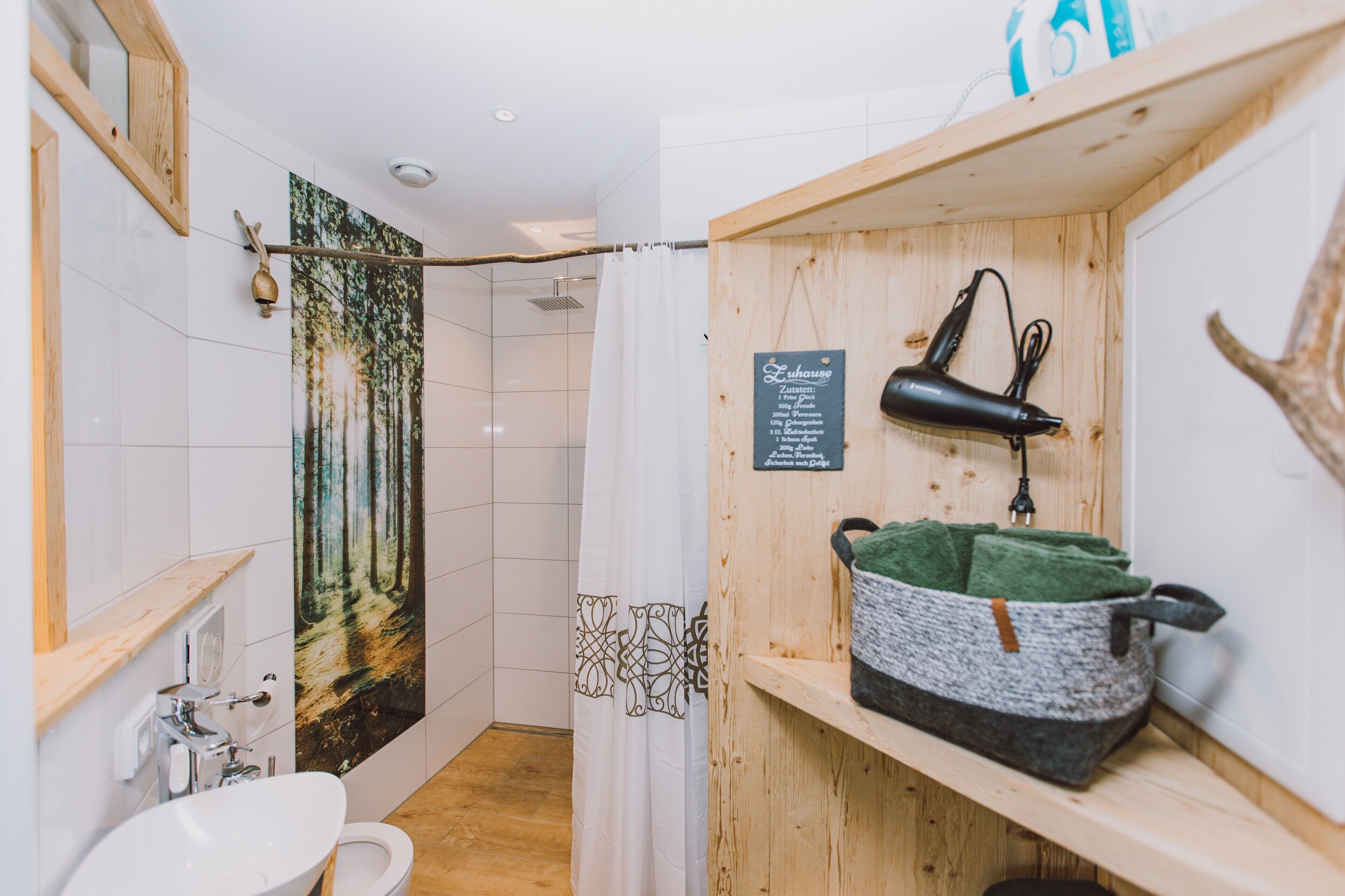 Une salle de bain avec un lavabo blanc, un miroir, un rideau de douche orné d’un motif de forêt et une étagère en bois sur laquelle reposent une serviette et un sac noir.