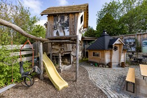 Eine Holzspielanlage mit Rutsche und Kletterelementen, umgeben von einem Kopfsteinpflasterweg und rustikalen Gebäuden.