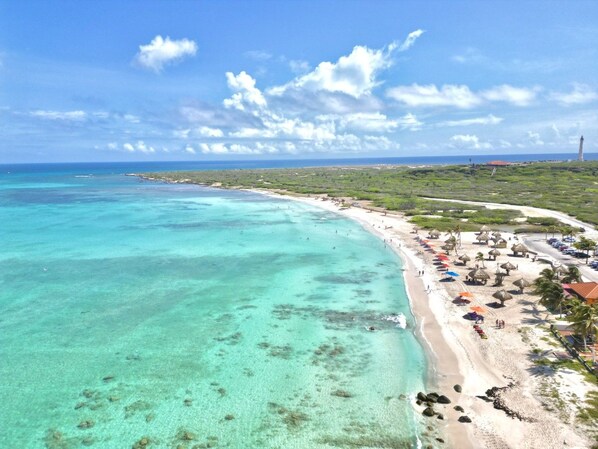 A coastal beach with clear turquoise waters, a sandy shore, and a row of thatched-roof structures along the beach.