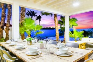 Un espace repas avec une longue table dressée pour un repas, entouré de plantes tropicales et offrant une vue sur le coucher de soleil.