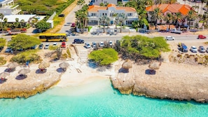 Vue aérienne d’une zone côtière avec une plage, des eaux turquoise et limpides, et un parking où sont stationnés plusieurs véhicules.