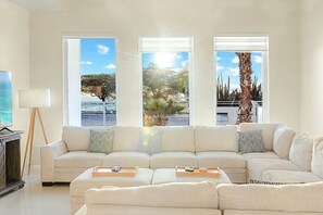 Un salon avec un canapé beige, une table basse et un lampadaire. Il y a trois grandes fenêtres qui offrent une vue sur l’extérieur.