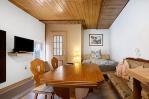 Une pièce comprenant une table à manger en bois, des chaises, un canapé, un lit recouvert d’une couverture à carreaux, une lampe et un téléviseur fixé au mur.