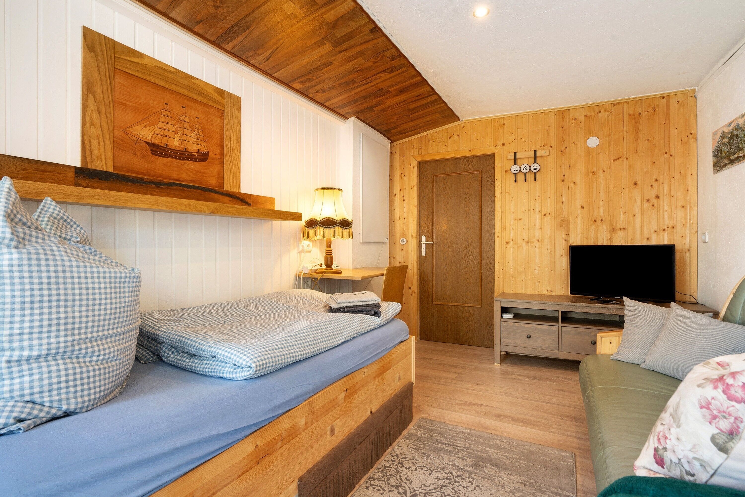Una habitación con una cama de madera, un escritorio de madera con una lámpara, un televisor y una puerta de madera.