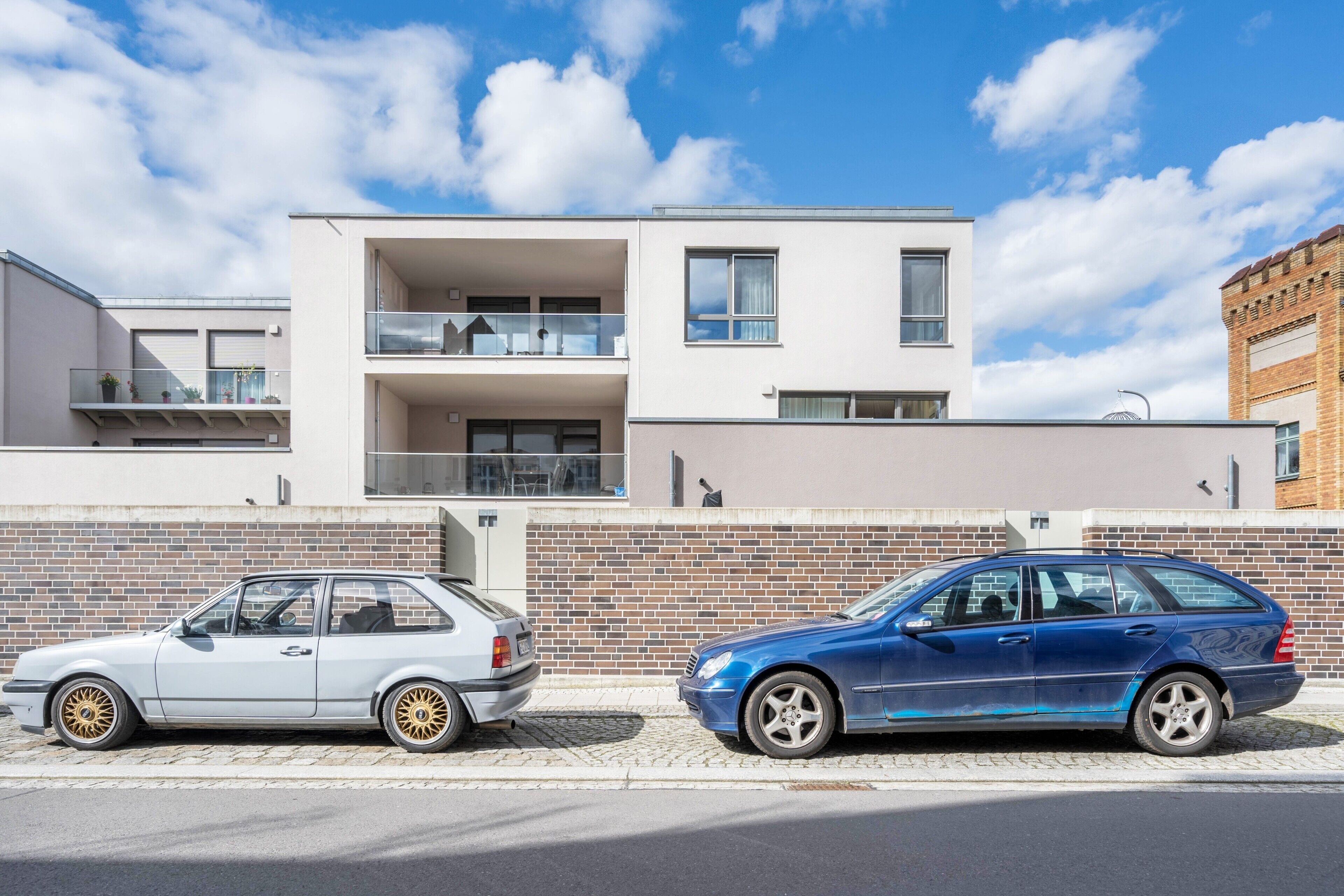Deux voitures sont garées devant un immeuble moderne à deux étages, doté d’une façade en briques et d’un balcon.
