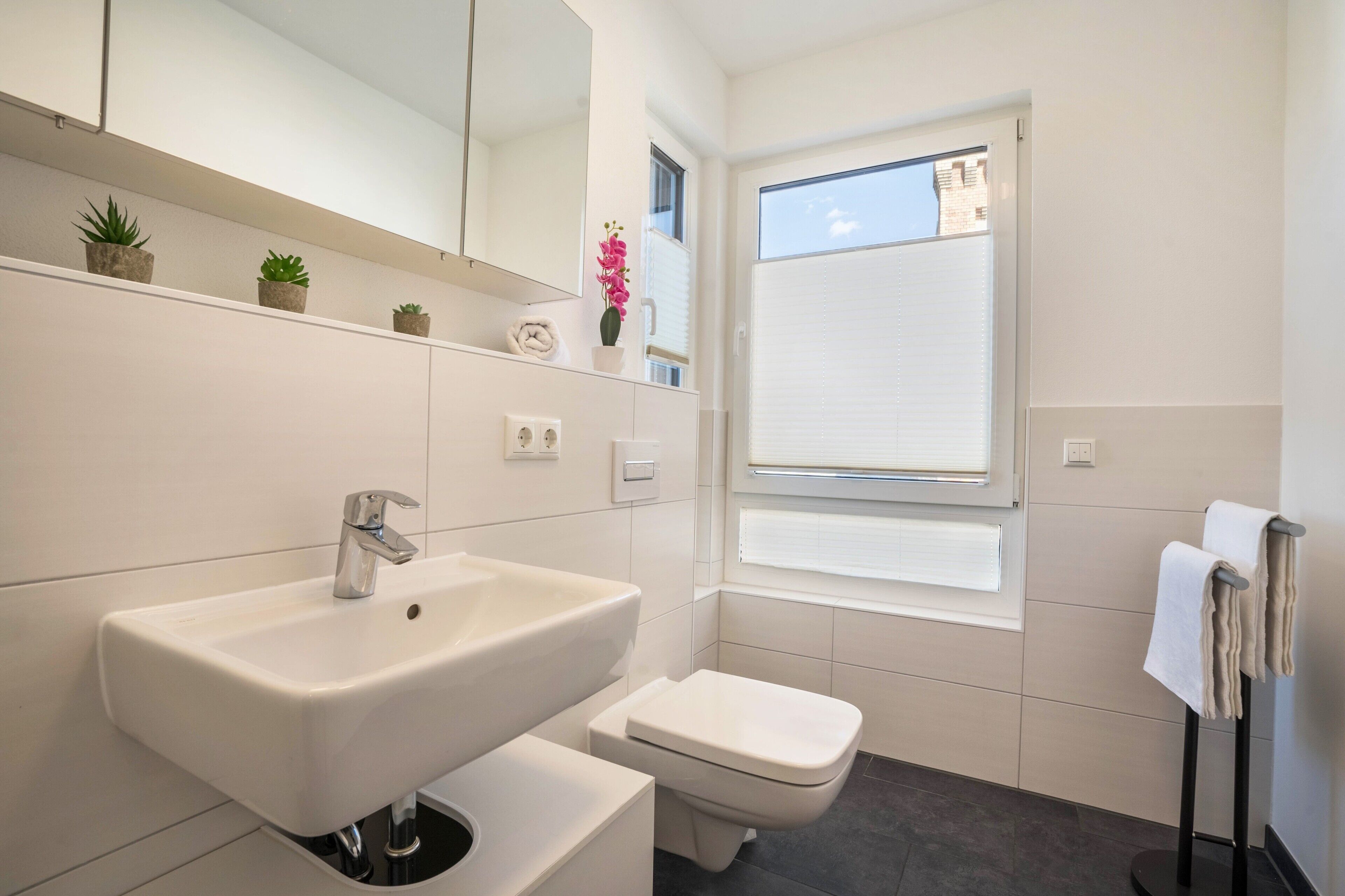 A modern bathroom with a white sink, toilet, and a window with blinds.