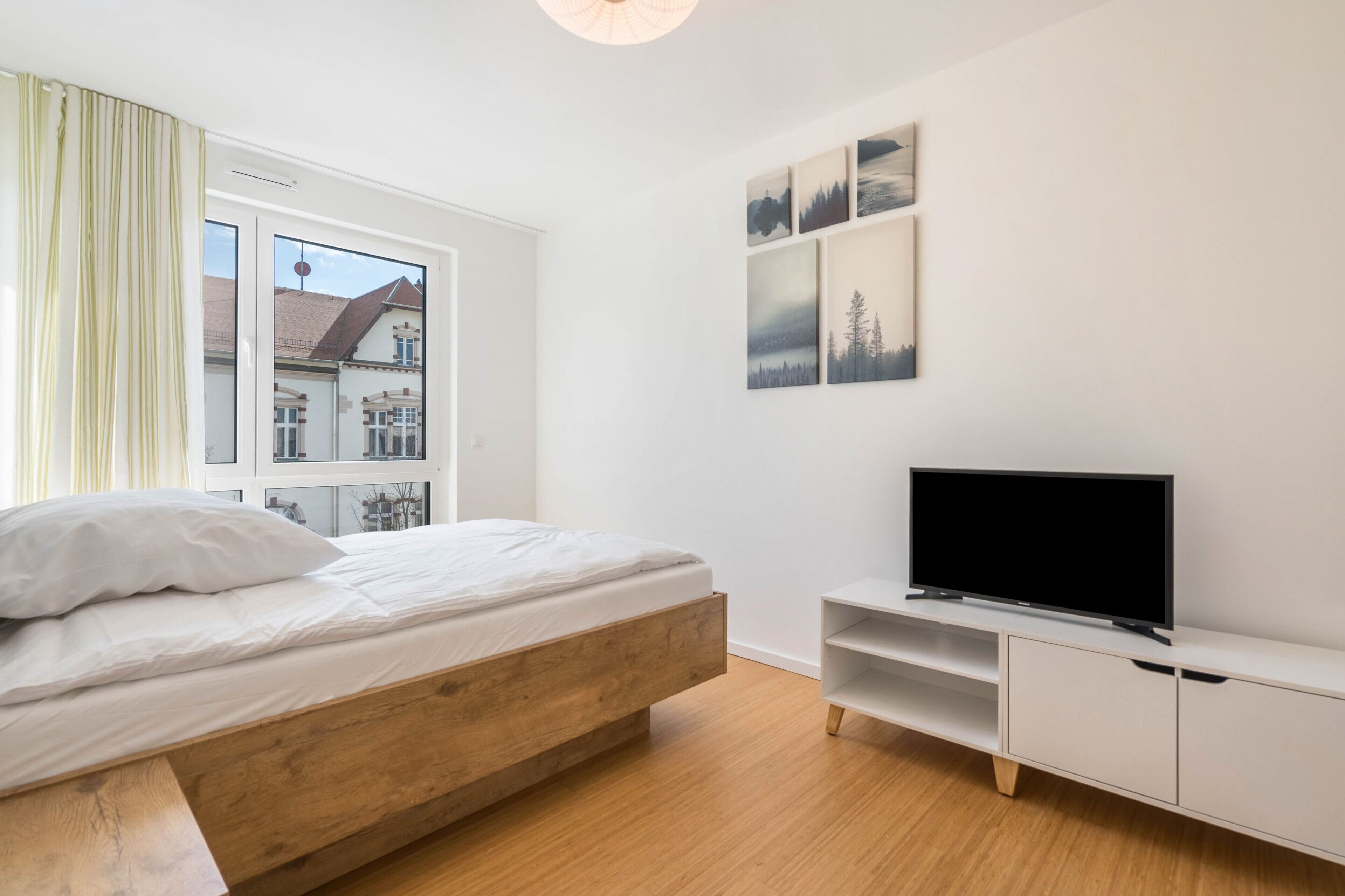 Un dormitorio con una cama de madera, un televisor sobre un soporte blanco y una ventana con cortinas.