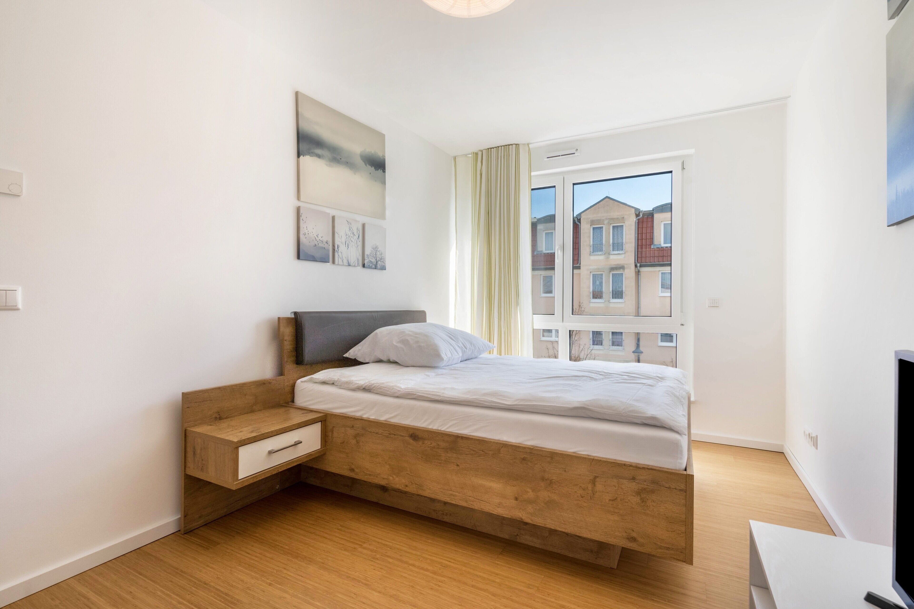 Ein Schlafzimmer mit einem hölzernen Bett, einem Nachttisch, einem Fernseher und einem Fenster mit Blick auf Gebäude.