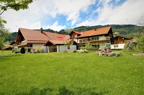 Un bâtiment de style chalet traditionnel, avec un toit recouvert de tuiles rouges, entouré d’une pelouse verdoyante et de plantes en fleurs.