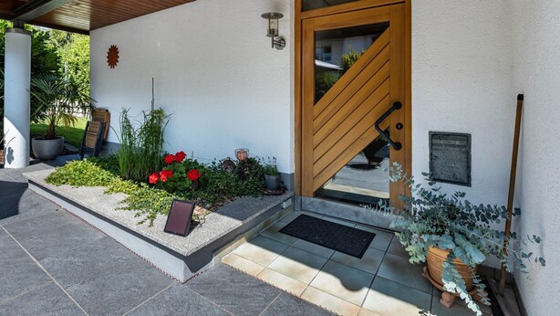 A modern building entrance with a wooden door, a small garden with red flowers, a potted plant, and a metal bench.