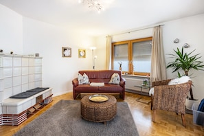 A living room with a sofa, ottoman, and wicker chair, a glass coffee table, a radiator, and a window with curtains.